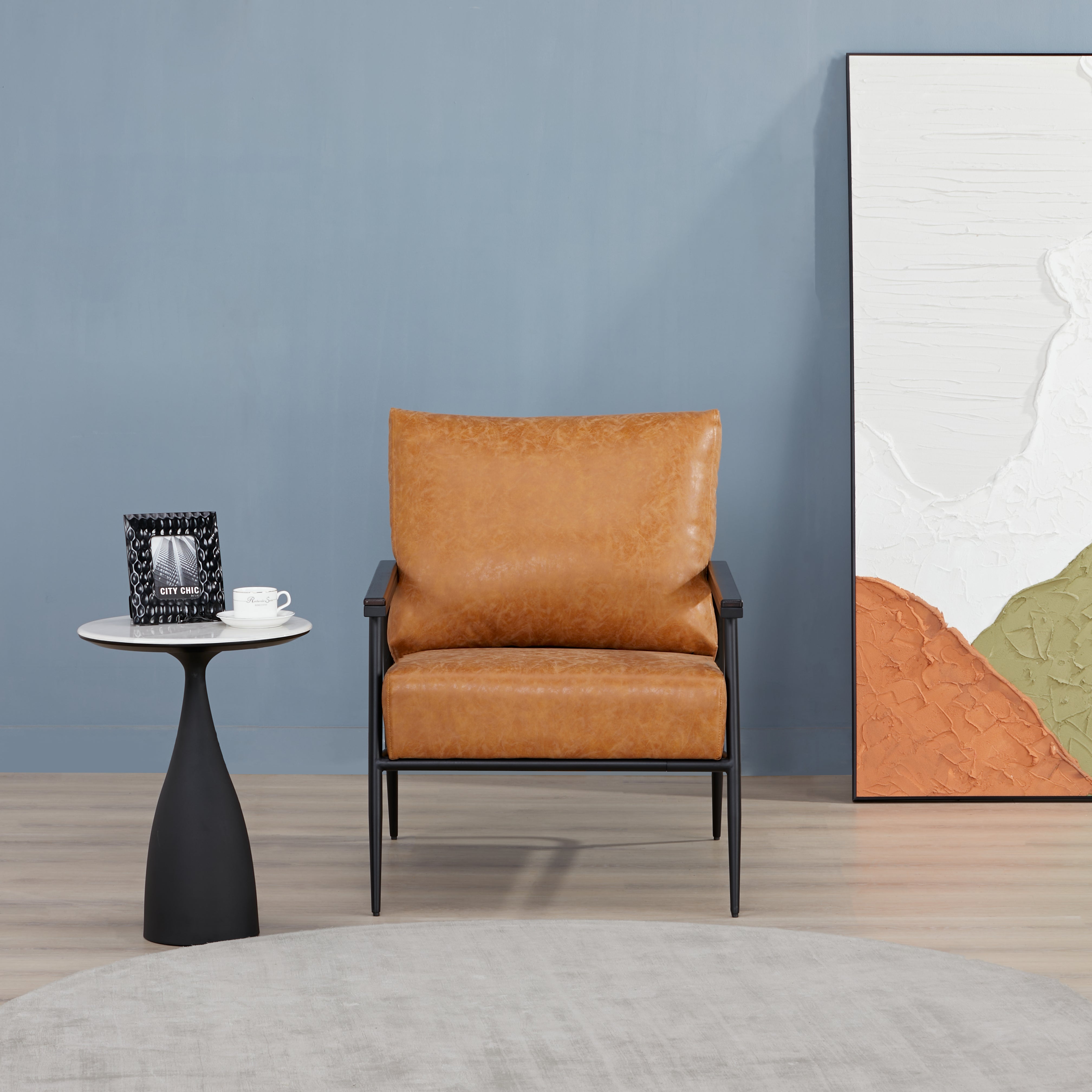 Parker Accent Chair in Faux Leather Camel displayed in a modern room with a side table, blue wall, abstract artwork, and camel faux leather upholstery